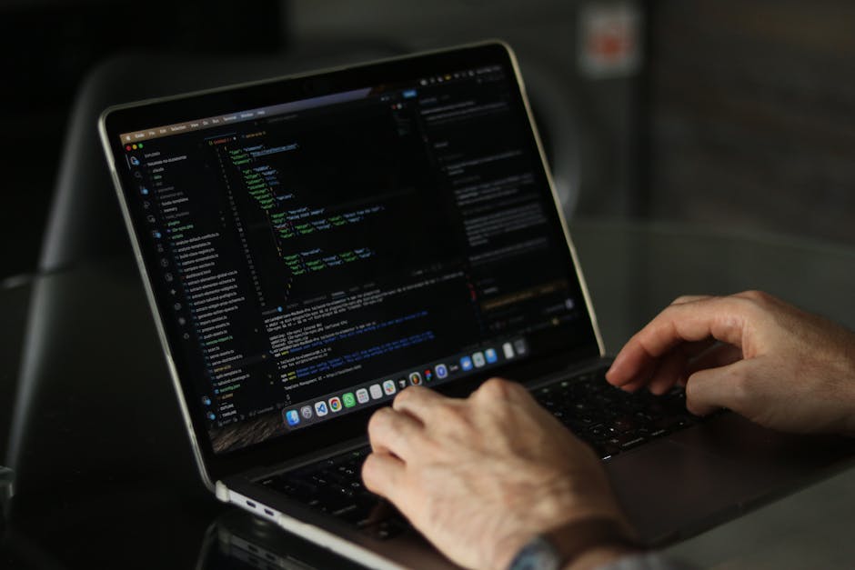 A close-up image showing hands working on a laptop with a dark screen displaying code in a programming environment. The user is seated at a desk, with the fingers positioned on the keyboard, possibly