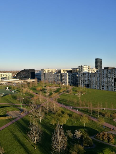 An expansive urban park with neatly maintained grassy areas and a network of winding pathways, bordered by young, leafless trees indicating early spring or late autumn, with some flowering trees in bl